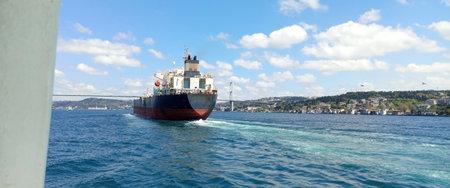 A cargo ship makes its way through the Bosphorus Strait in Istanbul, showcasing the vibrant blue water and clear sky with fluffy clouds. The bustling shoreline comes into view.の写真素材
