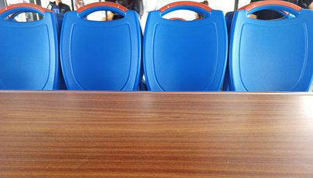 Four vibrant blue chairs are neatly aligned in front of a polished wooden table, creating a clean and inviting space. The background hints at a lively environment.の写真素材