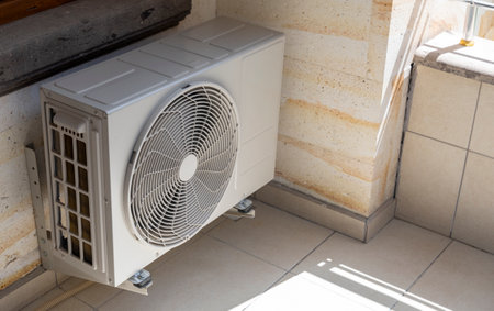 An air conditioning unit is mounted on the wall in a well-lit room. Sunlight filters through a window, casting patterns on the tiled floor.の写真素材