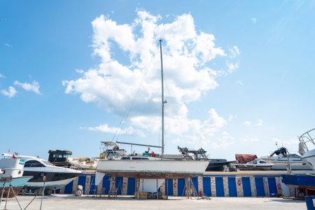 A sailboat rests on dry dock in a marina, surrounded by various vessels and bathed in bright...