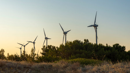 Wind turbines stand tall against a sunset sky, surrounded by a lush forest. The scene captures the beauty of renewable energy technology in harmony with nature.の写真素材