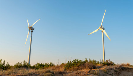 Two wind turbines tower over a green landscape under a clear blue sky, showing sustainable energy...