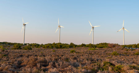 Tall wind turbines rotate slowly under a vibrant sky, surrounded by low shrubs and greenery. The...