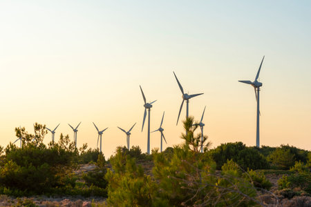 Golden sunlight bathes a row of wind turbines on a hillside, silhouetted against the soft hues of...