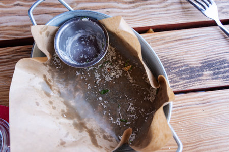 A metal serving dish with remnants of a meal sits on a wooden table. The plate is mostly empty, with traces of food and a small sauce cup.の写真素材
