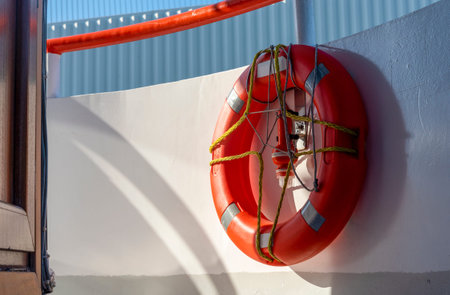 A bright orange lifebuoy is secured to a white wall on a boat. Sunshine illuminates the scene,...