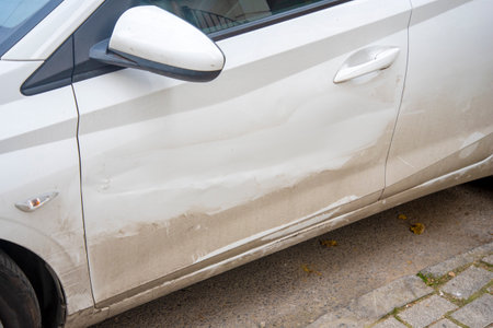 A white car shows visible scratches and dirt on its side while parked on a city street. The scene captures a typical urban environment during the day.の写真素材
