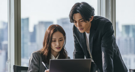 Two people are working together in a modern office space. They focus on a laptop while city buildings are visible through the large windows.の素材