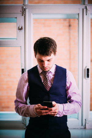 Handsome Caucasian Man In Business Attire Staying In Room Near Window And Holding Mobile Phoneの写真素材