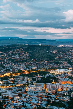 Tbilisi, Georgia. The Aerial Cityscape View Of Capital In Evening Illimination, The Modern City District Of High-Rise Buildings Foreground. Dramatic Blue Cloudy Sky Of Sunset Sunrise Over Hilly Area.の写真素材