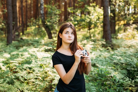 Happy Red-haired Caucasian Girl Young Woman Photographer Taking Pictures The Old Retro Vintage Film Camera In Summer Green Forest. Girl Dressed In A Black T-shirtの写真素材