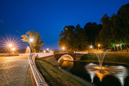 Gomel, Homiel, Belarus. Panorama Of Park Channel Flowing Into River Through Stone Bridge In City Park In Evening Or Night Illumination. Lighted Walkways, Greenwood Along Watercourse At Blue Hourの写真素材