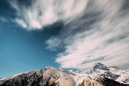 Stepantsminda, Georgia. Winter Cloudy Night Starry Sky With Glowing Stars Over Peak Of Mount Kazbek Covered With Snow. Beautiful Night Georgian Winter Landscapeの写真素材