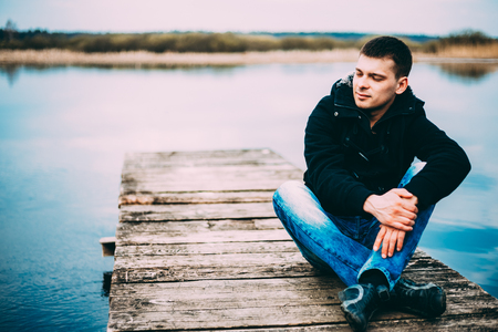 Young handsome man sitting on wooden pier, relaxing,  thinking, listening. Casual style - jeans, jacketの写真素材