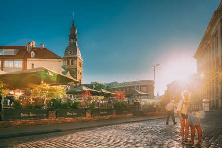 Riga, Latvia. Young Couple Tourists People Walking On Dome Square In Sunny Summer Day. Dome Cathedral On Background During Sunset.のeditorial素材