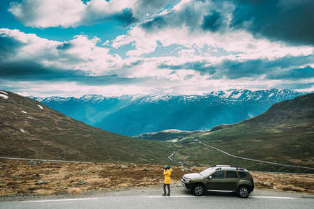 Aurlandsfjellet, Norway. Young Woman Tourist Traveler Photographer Taking Pictures Photos Near Parked Renault Duster Car. Aurlandsfjellet Scenic Route Road In Summer Norwegian Countryside Landscapeのeditorial素材