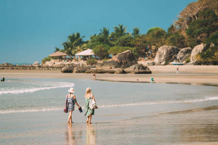 Canacona, Goa, India - February 16, 2020: Two Girls In Hats Walking Along Seashore On Palolem Beach With Shoes In Their Hands In Summer Sunny Dayのeditorial素材