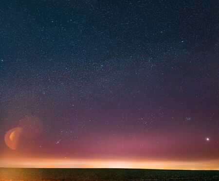 Night Starry Sky With Glowing Stars Above Countryside Landscape. Milky Way Galaxy And Rural Field Meadow In Early Spring. Bright Glow Of Planet Venus Among The Stars. Warm Lights Of Evening Sunset.の写真素材