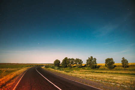 Blue Night Starry Sky Above Country Asphalt Road In Countryside And Green Meadow. Night View Of Natural Glowing Starsの写真素材
