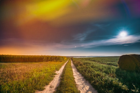 Night Starry Sky Above Country road through Field Meadow With Hay Bale After Harvest. Glowing Stars And Full Moon Above Rural Landscape. Colorful flare on sky. Agricultural Landscape under Starry Sky. Colored bright spots of light glow in the sky.の写真素材