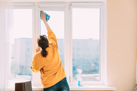 Woman Of Fifty In Yellow Sweater And Jeans Washes Dusty Window In Apartment. 50 Year Old Woman...
