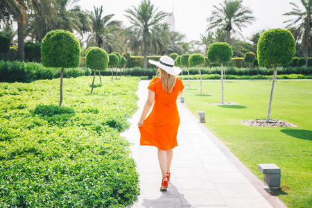 High quality photo. Young Caucasian Woman Lady In Red Dress And White Summer Hat Walking Through Trees In Zabeel Park In Sunny Summer Day. Back View. Landscape Design. Summertime. Summer Background. Resort In Dubaiの写真素材