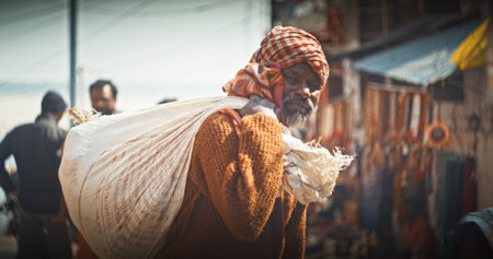 Varanasi, Uttar Pradesh, India. Old Indian man worker carries a heavy bag with a load.のeditorial素材