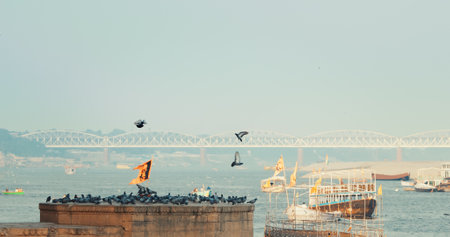 Varanasi, India. Bird Take Off from Sacred Ghat In Varanasi. Pigeons Fly Up In Slow Mo. Birds Flight. Orange Flags Fluttering In Slow Mo, Symbolizing Hanuman. Cinematic View On Ganga River. Many Pigeons Birds In Summer Day On Ghatの写真素材