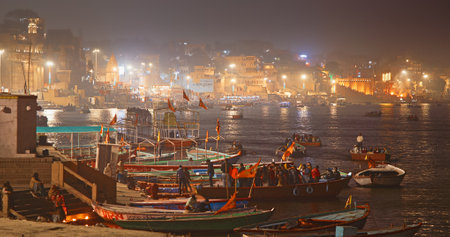 Varanasi, Uttar Pradesh, India. Many Boats are moored to the Ganga riverbank. Tourists boarding for an overnight boat ride on the Ganges River. Cityscape of Varanasi is visible - embankment illuminated in the twilight night lights. In the distance you canのeditorial素材