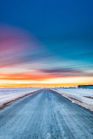 Asphalt Country Open Road Through Winter Snowy Fields And Meadows At Sunset Or Sunrise. Landscape In Belarus Or European Part Of Russiaの写真素材