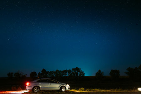 Car Sedan Parking On A Roadside Of Country Road On A Background Of Blue Sky At Winter Season. Country Asphalt Road In Countryside. Night View Of Natural Glowing Stars. Ground In Lights From Rear Red Car Lightの写真素材