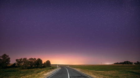 Night Starry Sky Above Country Road In Countryside And Green Field. Yellow City Lights On Background. Night View Of Natural Glowing Starsの写真素材