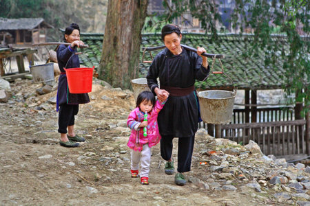 Zengchong village, Guizhou, China - April 11, 2010: Asian village, a Chinese woman goes fetch water, she carries pails on yoke, and leads by the hand of the child, a girl 3 years old, a child holding a toy, Zengchong Dong village, Guizhou, China - April 1のeditorial素材