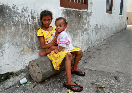 Zanzibar, Tanzania - February 16, 2008: Tanzania, Zanzibar island, Stone Town, a dark-skinned African girl 8 years old, sitting on a log near the house, she holds in her lap sister 2 years old, February 16, 2008.のeditorial素材