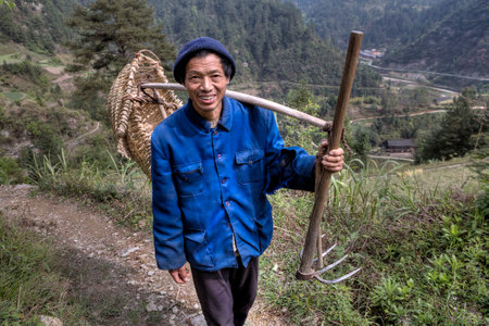 Langde Village, Guizhou, China - April 15, 2010: Chinese man, a villager, farmer, a peasant in a knitted cap and a blue work jacket, carries on the shoulder yoke with a wicker basket and a fork hoe.のeditorial素材