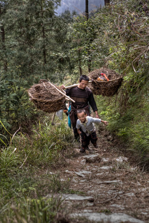 Langde Village, Guizhou, China - April 15, 2010: Chinese peasant farmer man carries the weight on your shoulder. Asian family farmer goes to work in  fields in highlands, boy runs next to his father.のeditorial素材