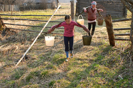 Lipovec village, Tver region, Russia - May 2, 2006: Girl carrying two heavy pails filled with water suspended from a shoulder yoke on her  farm.のeditorial素材