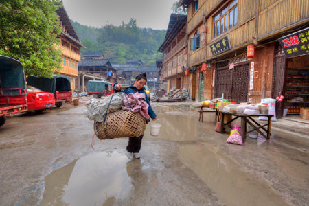Zhaoxing Dong Village, Guizhou, China -  April 8, 2010: Street tradeswoman with a yoke on his shoulder, carries a large wicker cage on street settlements of ethnic minorities passed wooden houses.のeditorial素材