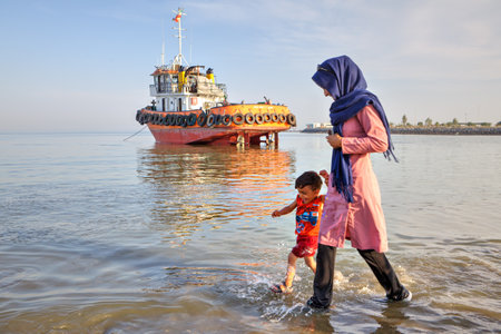 Bandar Abbas, Hormozgan Province, Iran - 16 april, 2017: A young woman walks along the shore of the Persian Gulf with her child, against the background of an orange towboat standing on the shallows.のeditorial素材