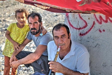 Bandar Abbas, Hormozgan Province, Iran - 16 april, 2017: Two adults man and one child girl relaxing outdoors in the shade of an awning, a man smokes a hookah.のeditorial素材