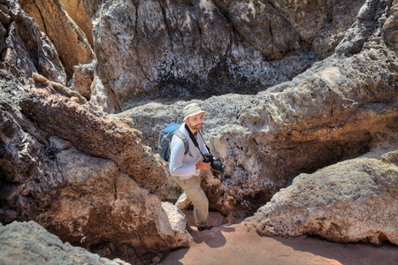 Off the beaten path travel destinations Hormuz Island Iran, one happy backpacker walks along a mountain path in bucket hat on his head, holding a camera in his hands, with a backpack behind him.の写真素材