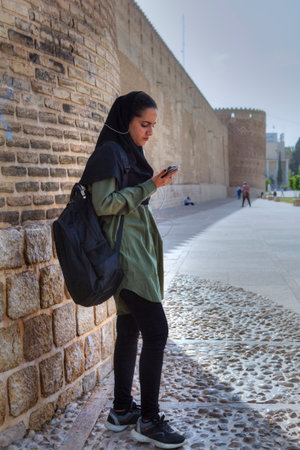 Fars Province, Shiraz, Iran - 19 april, 2017: A teenage girl in a Muslim hijab stands at the wall of Karim Khan Castle and listens to music from a smartphone using earphones.のeditorial素材