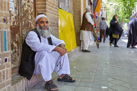 Yazd, Iran - April 22, 2017: An elderly bearded man in white Muslim clothes, fingering rosary, sitting on the threshold of a brick house, on a busy street.のeditorial素材