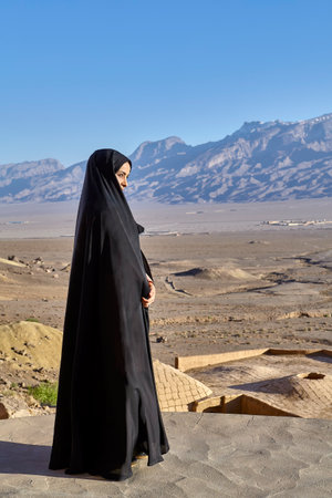 Yazd, Iran - April 21, 2017: One unknown Iranian Muslim girl, dressed in religious veil chador, stands on roof of  clay rural house, against backdrop of mountain landscape, on sunny evening.のeditorial素材