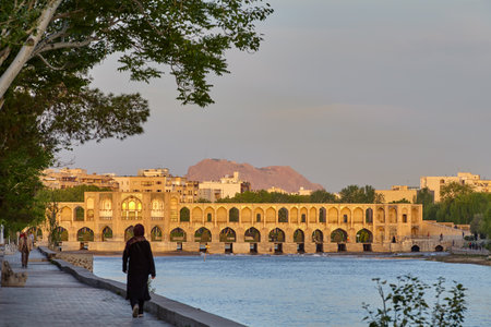 Isfahan, Iran - April 24, 2017: Townspeople walk along cobblestone road on embankment of Zayandeh River against backdrop of Khaju bridge, which is lit by rays of rising sun in early morning.のeditorial素材