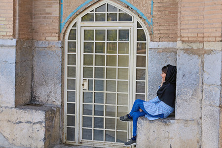 Isfahan, Iran - April 24, 2017: One young Iranian woman wearing a forced hijab, talking on the phone sitting on the stone architectural elements of the central pavilion Khaju bridge.のeditorial素材