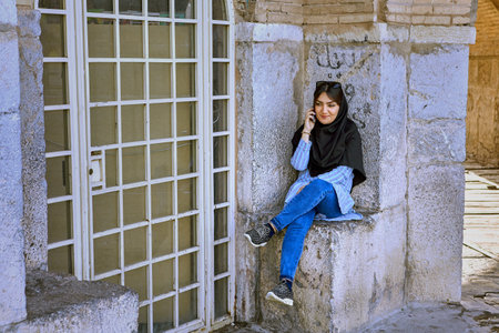 Isfahan, Iran - April 24, 2017: One young Iranian girl, wearing a forced hijab, is talking on the phone, sitting on the stone architectural element of the central pavilion of the Khaju bridge.のeditorial素材