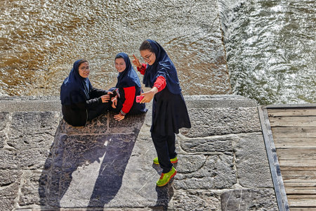 Isfahan, Iran - April 24, 2017: Three Iranian schoolgirls, dressed in an Islamic school uniform, take pictures of themselves near the Zayandeh river, using a pink smartphone.のeditorial素材