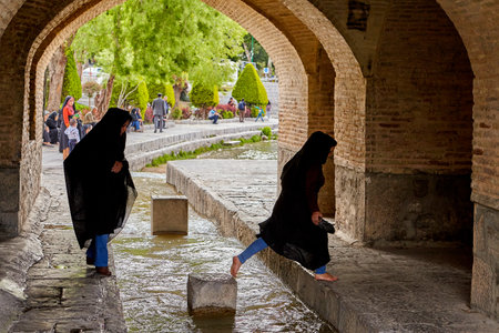 Isfahan, Iran - April 24, 2017: Two Iranian girls dressed in a black Muslim chador are crossing the stream of water under the bridge Allahverdi Khan, crossing the river Zayandeh.のeditorial素材