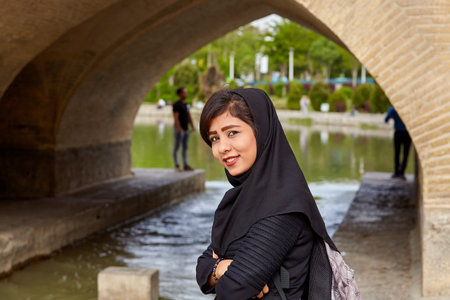 Isfahan, Iran - April 24, 2017: One young Iranian woman in black Muslim headscarf smiles when she observes that she is being photographed by tourist against background of Allahverdi Khan Bridge arch.のeditorial素材